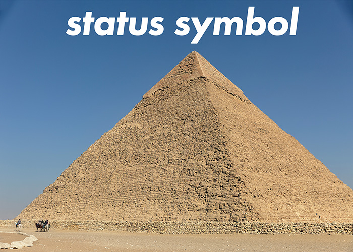 One of the great pyramids of Geyser against a mid-blue clear sky. A few people on horseback ride by its base, dwarfed in scale. Text above the pyramid reads 'status symbol'.